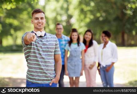 people, friendship and international concept - happy smiling man with headphones pointing at you and group of happy friends outdoors. group of happy international friends outdoors