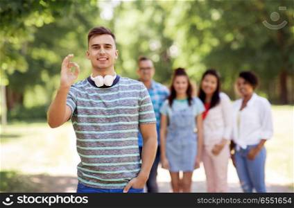people, friendship and international concept - happy smiling man with headphones showing ok hand sign and group of happy friends outdoors. group of happy international friends outdoors. group of happy international friends outdoors