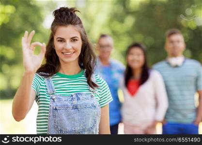 people, friendship and international concept - happy smiling woman showing ok hand sign and group of happy friends outdoors. group of happy international friends outdoors. group of happy international friends outdoors