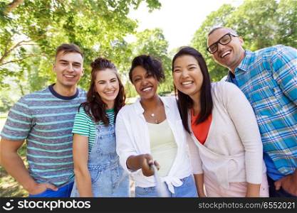 people, friendship and international concept - happy smiling young woman and group of happy friends taking picture by selfie stick outdoors. group of happy international friends taking selfie. group of happy international friends taking selfie