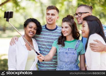 people, friendship and international concept - happy smiling young woman and group of happy friends taking picture by selfie stick in park. international friends taking selfie in park