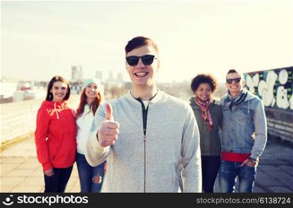 people, friendship and international concept - happy young man or teenage boy in front of his friends showing thumbs up on city street