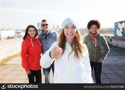 people, friendship and international concept - happy young woman or teenage girl pointing finger to you in front of her friends on city street
