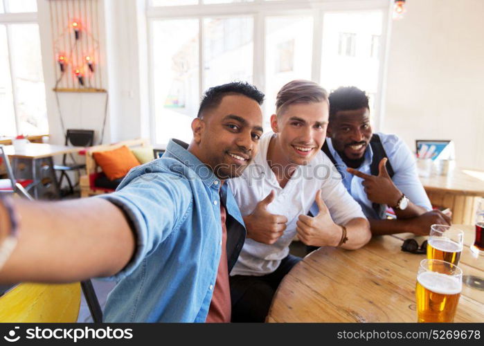 people, friendship and leisure concept - happy male friends drinking beer, taking selfie and showing thumbs up at bar or pub. friends taking selfie and drinking beer at bar