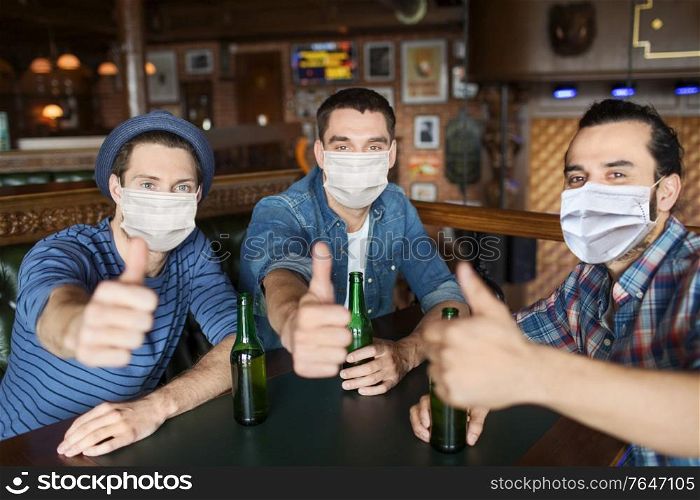 people, friendship and pandemic concept - male friends in face protective medical mask for protection from virus disease drinking bottled beer and showing thumbs up at bar or pub. male friends in masks drinking beer at bar or pub