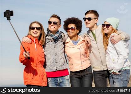 people, friendship and technology concept - group of smiling teenage friends taking picture with smartphone selfie stick outdoors. happy friends taking selfie by smartphone outdoors