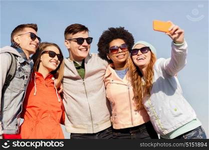 people, friendship and technology concept - group of smiling teenage friends taking selfie with smartphone outdoors. happy friends taking selfie by smartphone outdoors