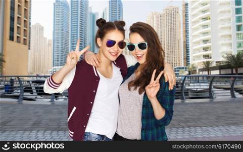 people, friendship, fashion, travel and tourism concept - happy smiling pretty teenage girls in sunglasses showing peace hand sign over dubai city street background