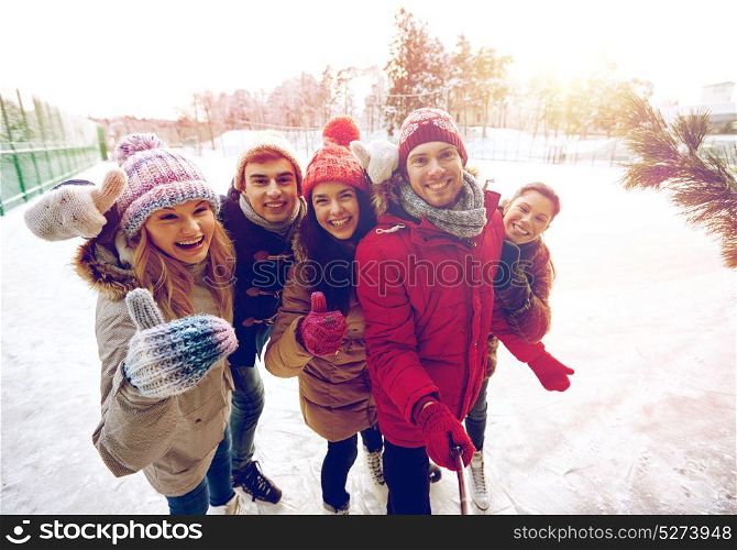 people, friendship, technology and leisure concept - happy friends taking picture with smartphone selfie stick and showing thumbs up on ice skating rink outdoors. happy friends with smartphone on ice skating rink