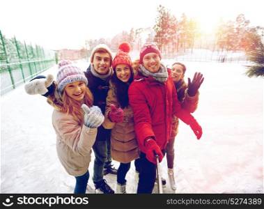 people, friendship, technology and leisure concept - happy friends taking picture with smartphone selfie stick and showing thumbs up on ice skating rink outdoors. happy friends with smartphone on ice skating rink