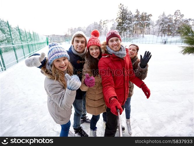 people, friendship, technology and leisure concept - happy friends taking picture with smartphone selfie stick and showing thumbs up on ice skating rink outdoors