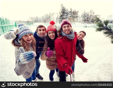 people, friendship, technology and leisure concept - happy friends taking picture with smartphone selfie stick and showing thumbs up on ice skating rink outdoors