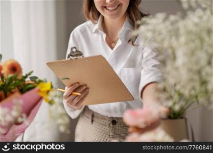 people, gardening and floral design concept - close up of happy smiling woman or floral artist with clipboard and flowers working at studio. happy woman with clipboard and flowers at studio