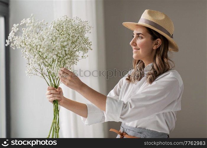 people, gardening and floral design concept - happy smiling woman or floral artist in straw hat holding bunch of gypsophila flowers at studio. happy woman holding gypsophila flowers at studio