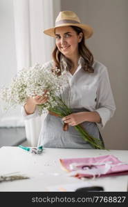 people, gardening and floral design concept - happy smiling woman or floral artist in straw hat with bunch of gypsophila flowers at studio. happy woman with gypsophila flowers at studio