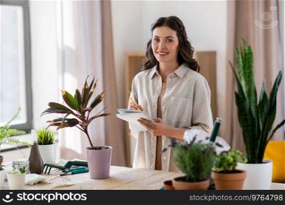 people, gardening and housework concept - happy smiling woman with notebook and pencil planting pot flowers at home. happy woman with notebook planting flowers at home
