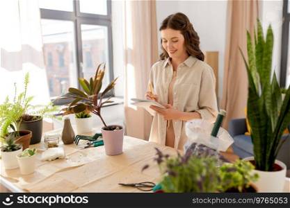 people, gardening and housework concept - happy smiling woman with notebook and pencil planting pot flowers at home. happy woman with notebook planting flowers at home