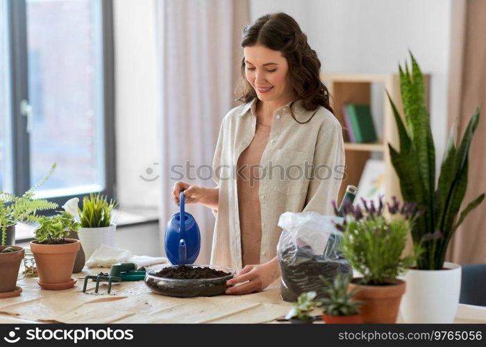 people, gardening and housework concept - happy woman in gloves with watering can and soil in glass vase planting pot flowers at home. happy woman planting pot flowers at home