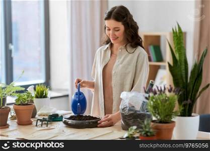people, gardening and housework concept - happy woman in gloves with watering can and soil in glass vase planting pot flowers at home. happy woman planting pot flowers at home