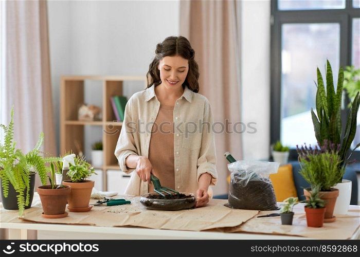 people, gardening and housework concept - happy woman with fork loosening soil in glass vase and planting flowers at home. happy woman planting pot flowers at home