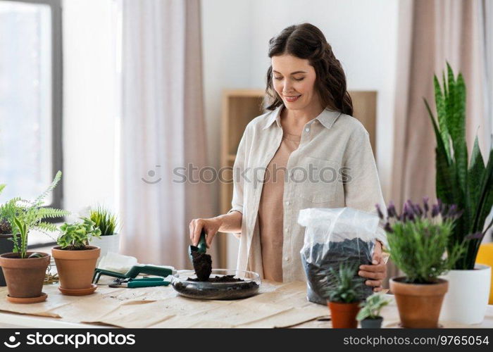 people, gardening and housework concept - happy woman with trowel and soil in bag planting flowers in glass vase at home. happy woman planting pot flowers at home