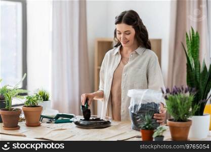 people, gardening and housework concept - happy woman with trowel and soil in bag planting flowers in glass vase at home. happy woman planting pot flowers at home