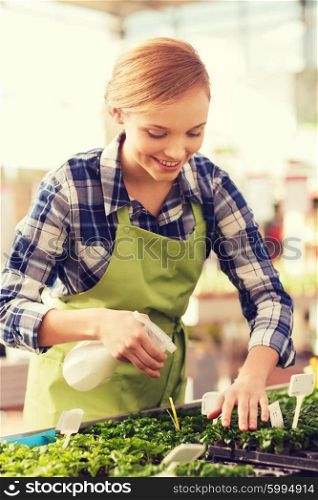 people, gardening and profession concept - happy woman or gardener with sprayer and seedling in greenhouse