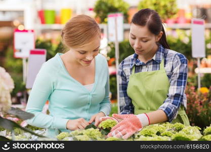 people, gardening, shopping, sale and consumerism concept - happy gardener helping woman with choosing flowers in greenhouse