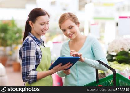 people, gardening, shopping, sale and consumerism concept - happy gardener with tablet pc helping woman with choosing flowers in greenhouse
