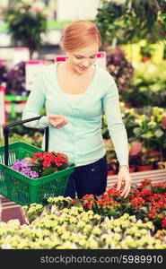 people, gardening, shopping, sale and consumerism concept - happy woman with basket choosing and buying flowers in greenhouse