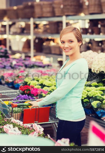 people, gardening, shopping, sale and consumerism concept - happy woman with trolley buying flowers at greenhouse or shop