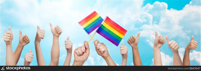 people, gay pride, gesture and homosexual concept - human hands showing thumbs up and holding rainbow flags over blue sky background