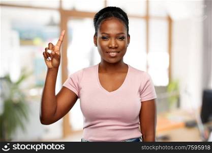 people, gesture and corporate concept - happy african american young woman pointing finger up over office background. happy african woman pointing finger up at office