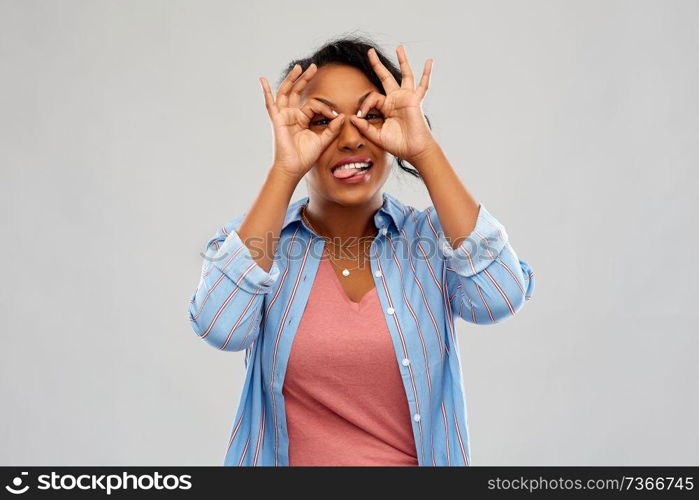 people, gesture and fun concept - happy african american young woman looking through finger glasses and sticking tongue out over grey background. african woman looking through finger glasses