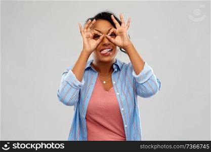people, gesture and fun concept - happy african american young woman looking through finger glasses and sticking tongue out over grey background. african woman looking through finger glasses