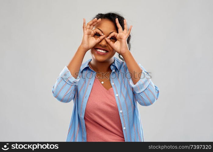 people, gesture and portrait concept - happy african american young woman looking through finger glasses over grey background. african woman looking through finger glasses