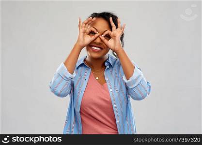 people, gesture and portrait concept - happy african american young woman looking through finger glasses over grey background. african woman looking through finger glasses