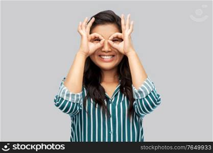 people, gesture and portrait concept - smiling young asian woman looking through finger glasses over grey background. smiling asian woman looking through finger glasses