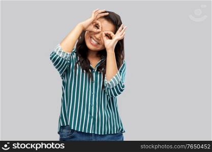 people, gesture and portrait concept - smiling young asian woman looking through finger glasses over grey background. smiling asian woman looking through finger glasses