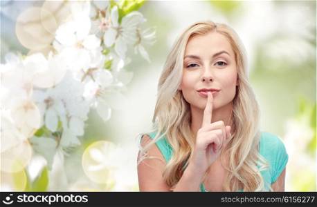 people, gesture and secret concept - beautiful young woman holding finger at her lips over natural spring background