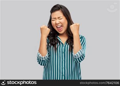 people, gesture and winning concept - happy asian young woman celebrating success over grey background. happy asian woman celebrating success over grey