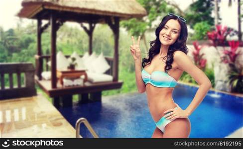 people, gesture, summer holidays, travel and tourism concept- happy woman in bikini swimsuit showing victory hand sign over hotel resort background