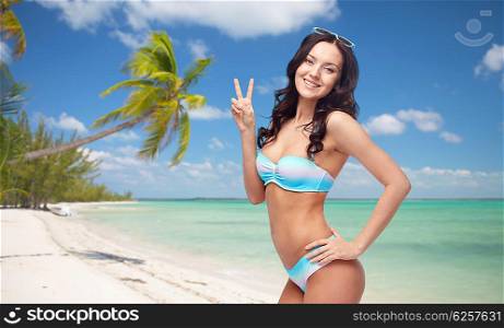 people, gesture, tourism, travel and summer concept - happy woman in bikini swimsuit showing victory hand sign over tropical beach background