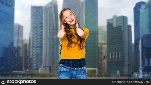 people, gesture, travel, tourism and fashion concept - happy young woman or teen girl in casual clothes showing thumbs up over city buildings background