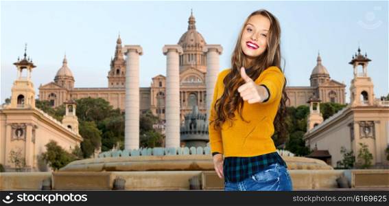 people, gesture, travel, tourism and fashion concept - happy young woman or teen girl in casual clothes showing thumbs up over national museum of barcelona background