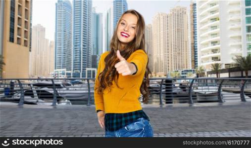 people, gesture, travel, tourism and fashion concept - happy young woman or teen girl in casual clothes showing thumbs up over dubai city street background
