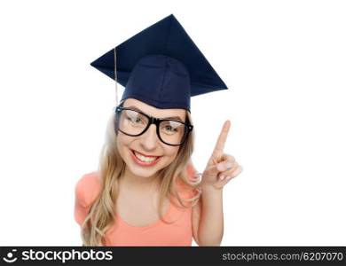 people, graduation and education concept - smiling young student woman in mortarboard and eyeglasses pointing finger up