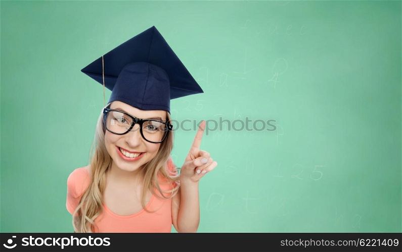 people, graduation and education concept - smiling young student woman in mortarboard and eyeglasses pointing finger up over green school chalk board background