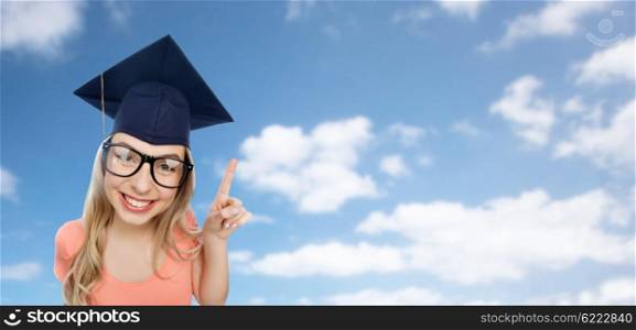people, graduation and education concept - smiling young student woman in mortarboard and eyeglasses pointing finger up over blue sky and clouds background