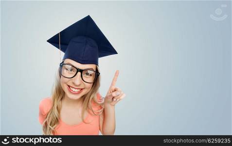 people, graduation and education concept - smiling young student woman in mortarboard and eyeglasses pointing finger up over gray background